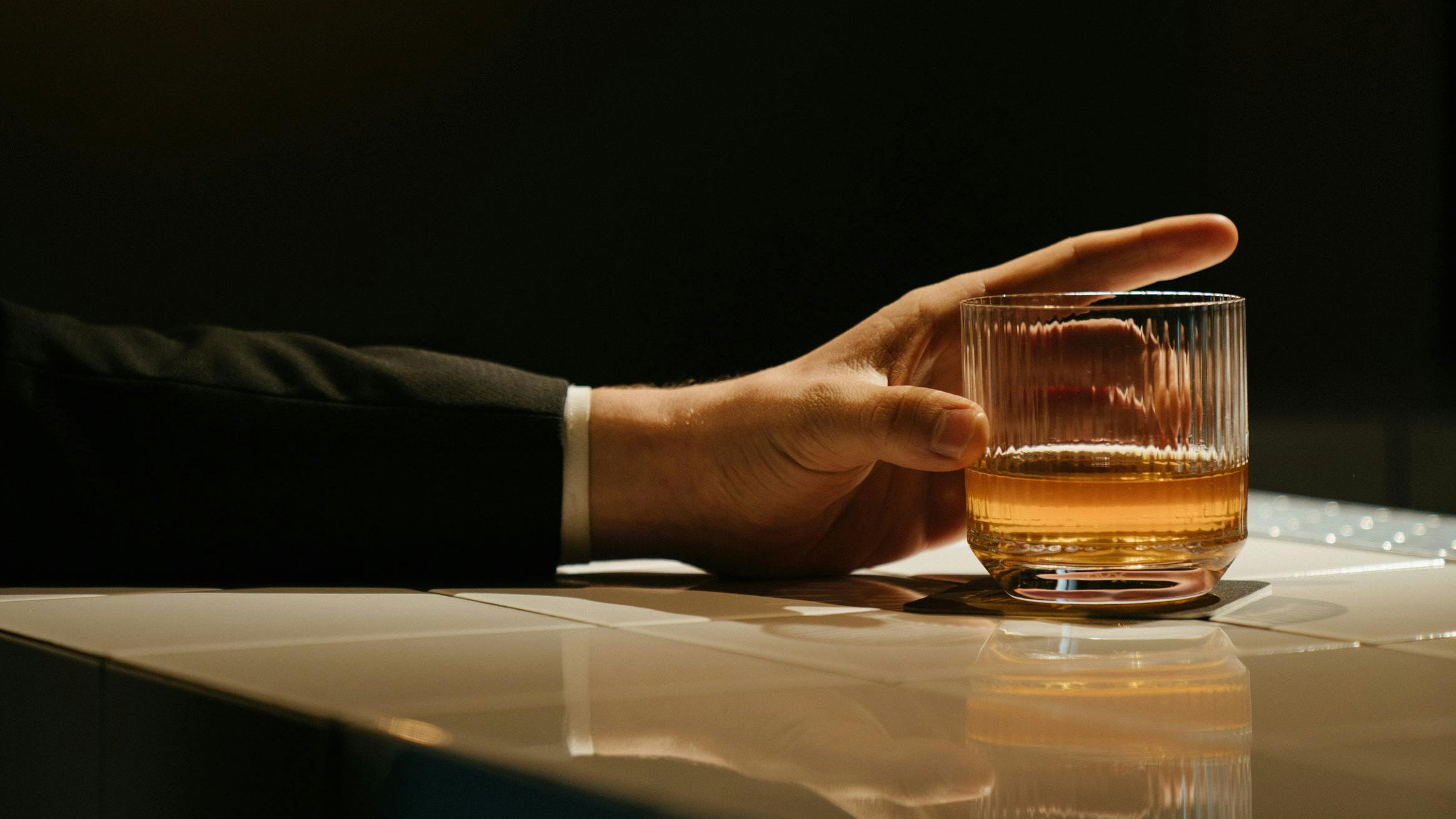 Nahaufnahme einer Hand im Anzug, die ein Glas mit bernsteinfarbenem Alkohol auf einem Tresen hält – symbolisches Bild für Erektionsstörung durch Alkohol.