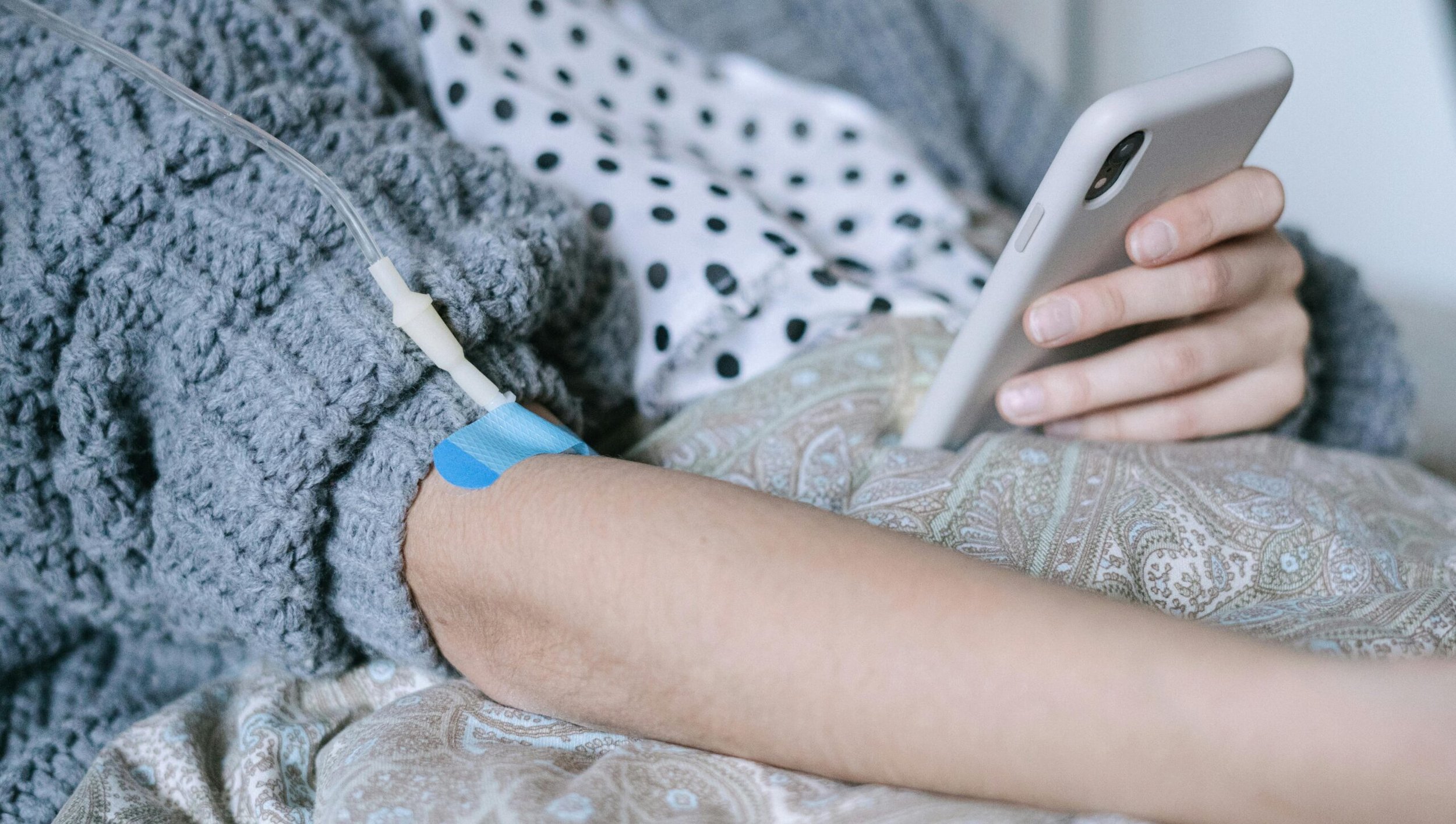 Chemotherapie Haarausfall: Das Bild zeigt eine Person, die während einer Chemotherapie auf einem Bett liegt. Der Arm der Person ist mit einem blauen Pflaster fixiert, durch das ein Infusionszugang gelegt wurde. Die Person trägt bequeme Kleidung und hält ein Smartphone in der Hand, was darauf hindeutet, dass sie versucht, die Zeit während der Therapie zu überbrücken.