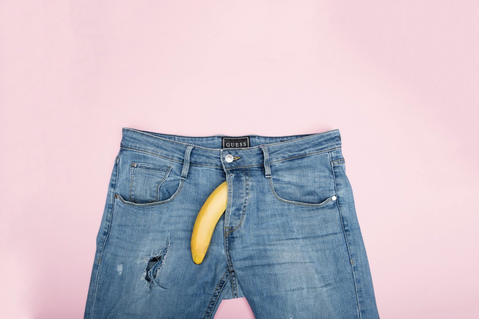 Eine Jeanshose mit einer Banane, die aus dem Hosenschlitz herausragt und nach unten zeigt, vor einem rosafarbenen Hintergrund. Es symbolisiert erektile Dysfunktion.