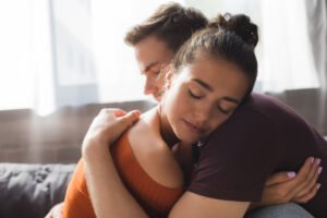 Ein Mann und eine Frau sitzen eng umschlungen auf einer Couch. Beide haben die Augen geschlossen, und die Umarmung wirkt beruhigend und unterstützend. Die Frau trägt ein orangefarbenes Oberteil, der Mann ein dunkles Shirt. Im Hintergrund ist ein lichtdurchflutetes Fenster zu sehen. Ein Teil von Erektionsstörung was kann frau tun.