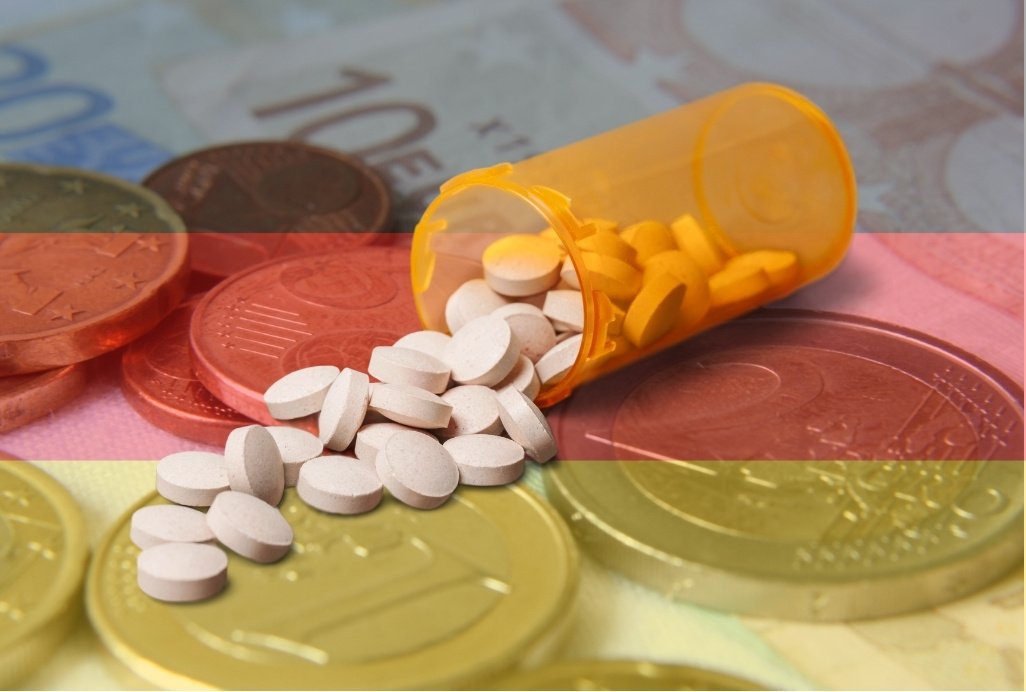 Euromünzen und Scheine mit einer transparent gehaltenen Deutschland-Flagge im Vordergrund. Mittig ist ohne Transparenz eine ausgeschüttete Medikamentendose mit weißen Tabletten zu sehen.