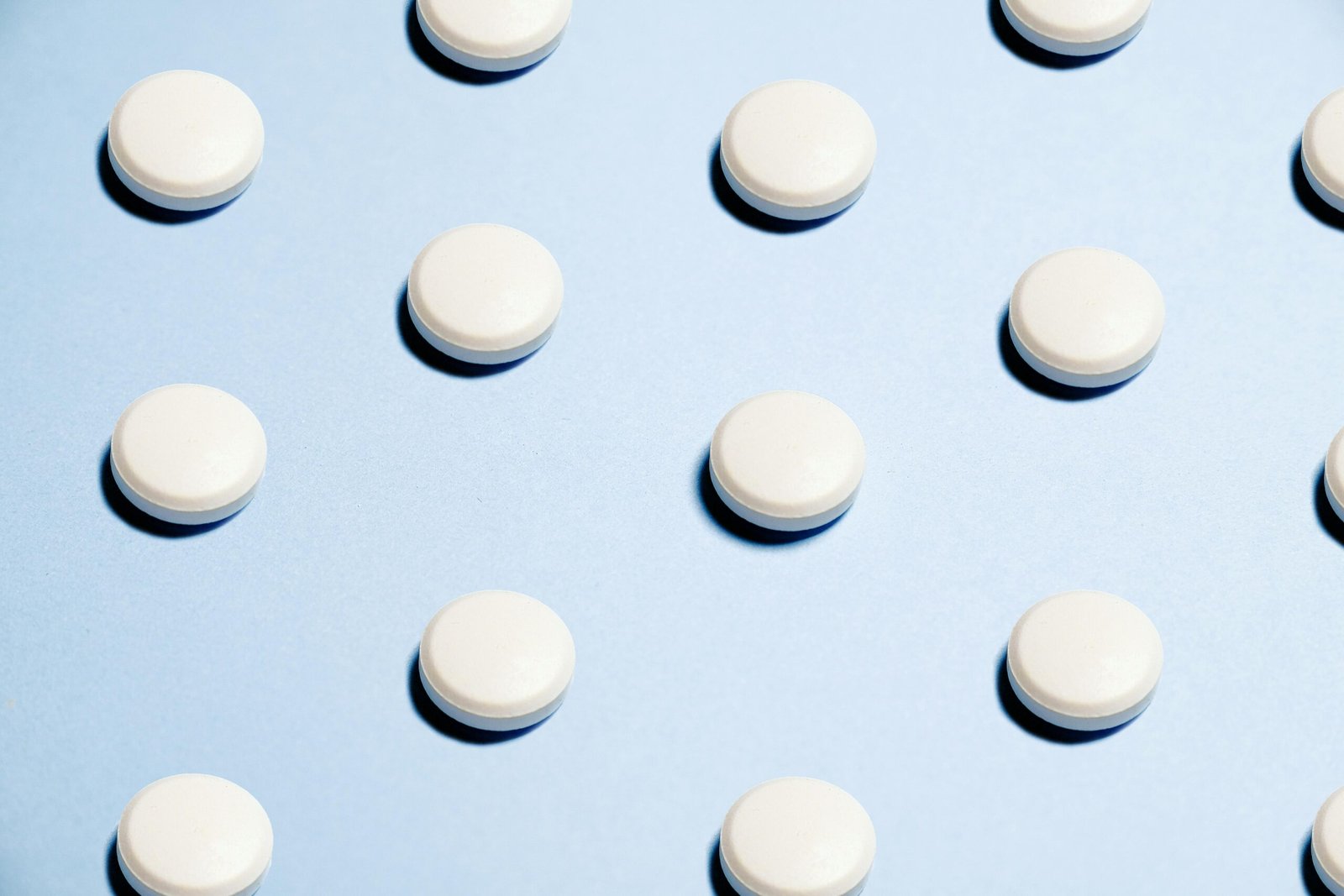 Mehrere weiße, runde Tabletten als Zeichen für Finasterid sind gleichmäßig auf einem hellblauen Hintergrund angeordnet. Jede Tablette wirft einen kleinen Schatten.