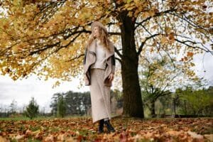 Eine Frau mit lockigem Haar steht unter einem Baum mit goldenen Herbstblättern, passend zum Thema Haarausfall Winter und Herbst, während der Boden mit herabgefallenen Blättern bedeckt ist.