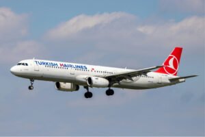 Das Bild zeigt ein Flugzeug von Turkish Airlines, das vor Wolken in der Luft fliegt. Auf dem Flugzeug steht Turkish Hairlines, wobei das H von Hairlines in dunklem Rot hervorgehoben ist. Es ist als Symbolbild für Haartransplantation Türkei gedacht.