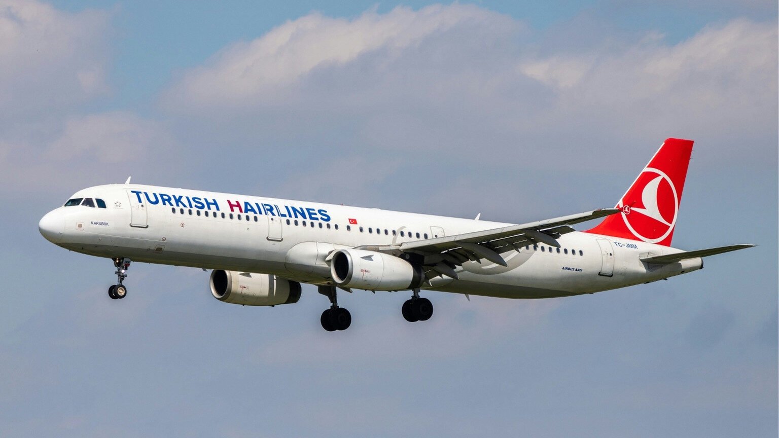 Das Bild zeigt ein Flugzeug von Turkish Airlines, das vor Wolken in der Luft fliegt. Auf dem Flugzeug steht Turkish Hairlines, wobei das H von Hairlines in dunklem Rot hervorgehoben ist. Es ist als Symbolbild für Haartransplantation Türkei gedacht.