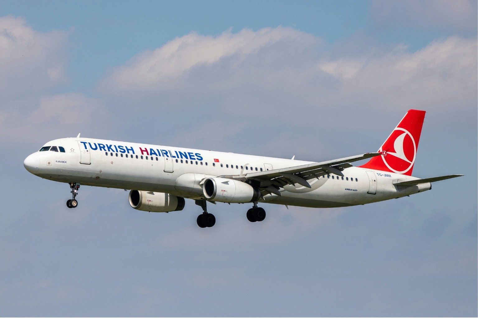 Das Bild zeigt ein Flugzeug von Turkish Airlines, das vor Wolken in der Luft fliegt. Auf dem Flugzeug steht Turkish Hairlines, wobei das H von Hairlines in dunklem Rot hervorgehoben ist. Es ist als Symbolbild für Haartransplantation Türkei gedacht.