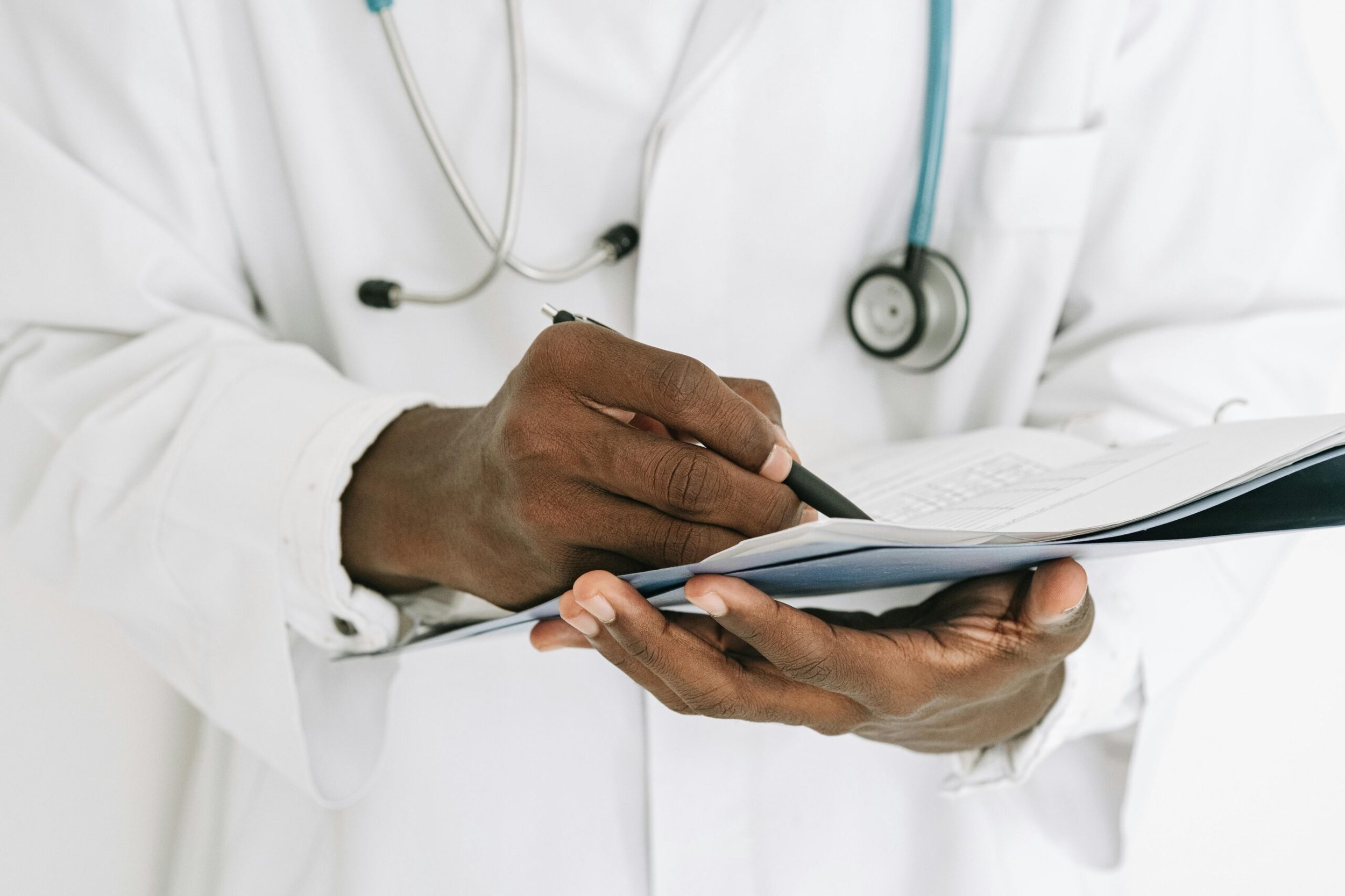 Libidoverlust Diagnose: Ein Arzt in einem weißen Kittel schreibt etwas auf ein Klemmbrett. Um seinen Hals hängt ein Stethoskop, und er scheint konzentriert zu arbeiten. Nur der Oberkörper und die Hände sind sichtbar. Es ist ein Symbolbild für die Anamnese zur Diagnose von Libidoverlust.