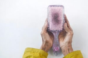 Plötzlicher Haarausfall im hohen Alter: Eine ältere Person hält eine Haarbürste mit vielen ausgefallenen Haaren. Die faltigen Hände und der gelbe Ärmel deuten auf das hohe Alter hin. Die Bürste hat ein glitzerndes, lila Design.