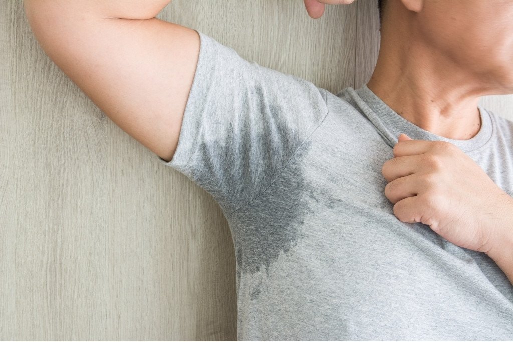 Person, die starkes Schwitzen unter den Achseln erlebt, hebt den rechten Arm vor einem holzernen Hintergrund. Das hellgraue T-Shirt hat sichtliche nasse Flecken.